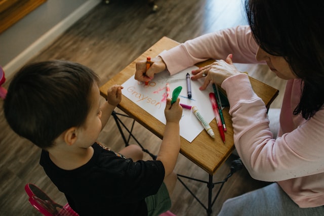 A home daycare worker is assisting a young boy in drawing on a piece of paper.