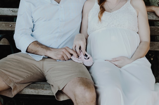 A pregnant couple sitting on a bench holding baby shoes. 